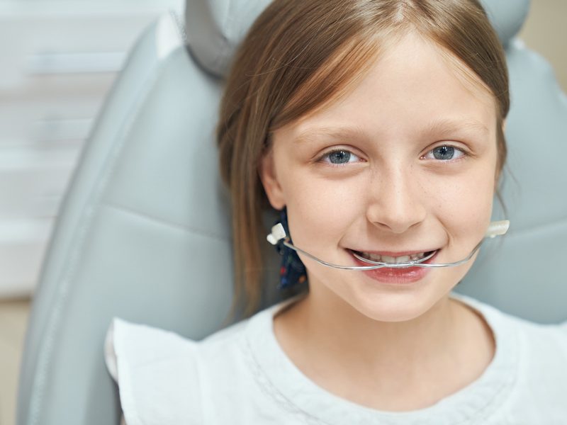 Happy cute little girl with beautiful blue eyes sitting in dental chair with orthodontic braces for improving bite. Young pretty lady taking care about teeth in professional clinic